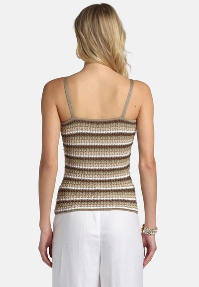 Femme aux cheveux blonds portant un débardeur en crochet rayé marron, blanc et beige et un pantalon blanc, vue de dos.