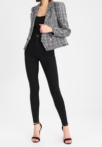 Žena v černých vysokých skinny džínách, černém topu, černo-bílé tweedové saku s roztrhanými okraji a černých otevřených lodičkách.