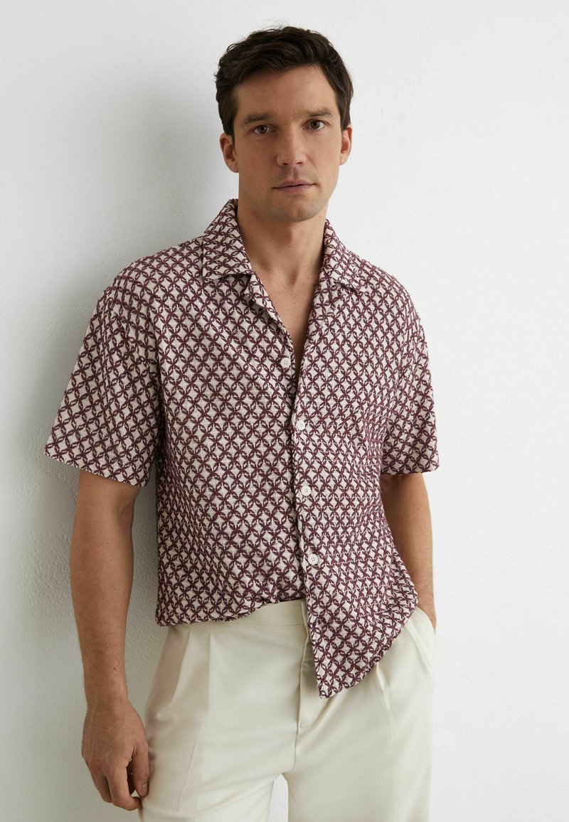 Uomo con capelli scuri che indossa una camicia a maniche corte color bordeaux e bianca con motivi, infilata in pantaloni crema a vita alta.