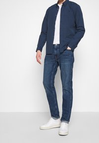 Camisa de botones azul oscuro sobre una camiseta blanca, combinada con unos vaqueros oscuros ajustados y zapatillas blancas. Tela suave y diseño clásico.