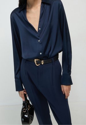 Frau trägt eine dunkelblaue Satinbluse mit Knöpfen, passende hoch taillierte Hose, schwarzen Gürtel mit goldener Schnalle, hält eine kleine schwarze Lackhandtasche.