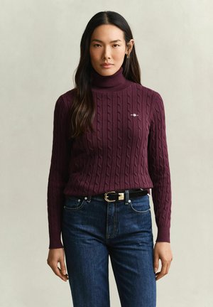 Frau mit langen dunklen Haaren, die einen bordeauxfarbenen, kabelgestrickten Rollkragenpullover und blaue Jeans mit einem schwarzen Gürtel trägt und vor einem einfarbigen Hintergrund steht.