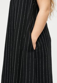 Το χέρι μιας γυναίκας στην τσέπη μιας μαύρης φούστας με κάθετες λευκές λεπτές ρίγες, που δείχνει μέρος του βραχίονα και κυματιστά ξανθά μαλλιά πάνω σε λευκό φόντο.