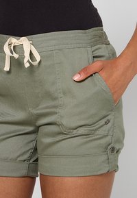 Personne portant un short à cordon de serrage vert olive avec ourlets retroussés et poches, la main partiellement dans la poche droite.