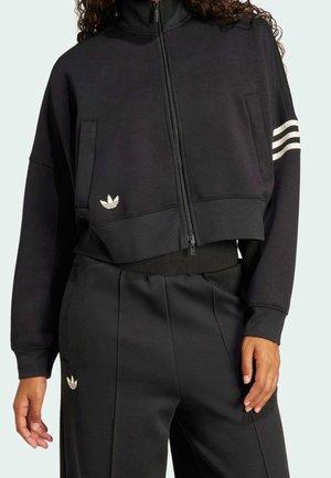 Personne portant une veste courte noire zippée avec des bandes blanches sur une manche et un pantalon noir assorti avec des logos blancs.