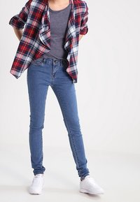 Rutigt röd och blå skjorta över en grå topp, i kombination med åtsittande blå jeans och vita sneakers. Slät tyg och avslappnad passform.