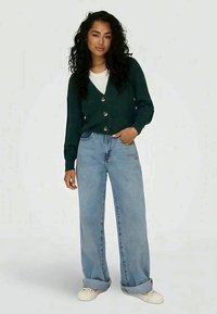 Cardigã de malha verde escuro com três botões, usado sobre uma camisa branca; combinado com jeans de perna larga azul claro e ténis brancos.