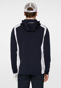 Marinblå huvtröja med vita sidokanter, med huva, långa ärmar och mjuk textur. På baksidan visas varumärkets logotyp vid nacken.