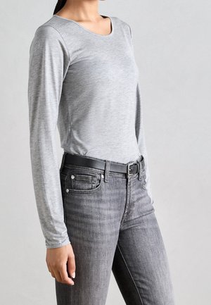 Osoba v dlouhém světle šedém tričku zasunutém do vybledlých černých džín s vysokým pasem a černým pásem, na jednobarevném šedém pozadí.