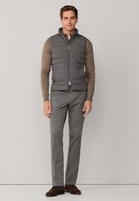 Grijze donsvest met een hoge kraag, gedragen over een bruin shirt met lange mouwen. Grijze broek met een slanke pasvorm, aangevuld met bruine schoenen.