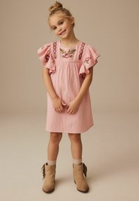 Rosenfarbiges Baumwollkleid mit Rüschenärmeln, floralem besticktem Oberteil und strukturierter Oberfläche. Kombiniert mit hellbraunen Knöchelstiefeln und beigen Socken.