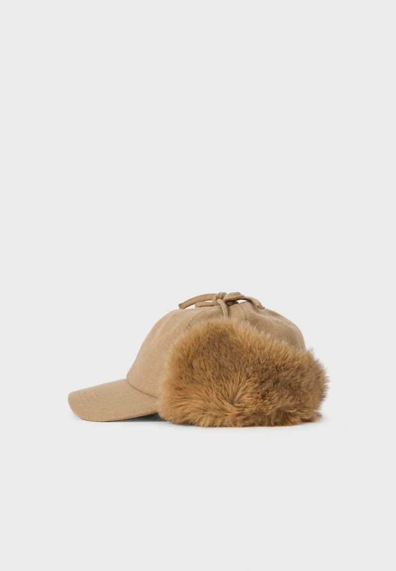 Beige winter cap with a short brim and large fluffy fur ear flaps against a plain white background.