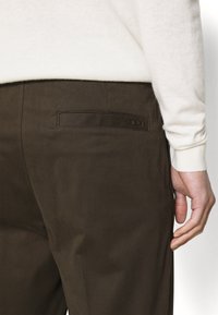 Mörkbruna byxor med en slät textur, som har en bakficka med en subtil läderdetalj och ett litet präglat nummer "1903."