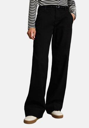 Person trägt schwarze Hose mit weitem Bein und schwarzem Gürtel, ein schwarz-weiß gestreiftes Langarmshirt und weiße Sneakers mit braunen Sohlen.