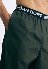 Calções verdes escuros de material leve, com uma larga cintura preta com "BJÖRN BORG" em letras brancas.