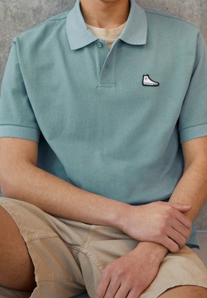 Polo bleu clair à manches courtes, avec un petit logo de baskets noir et blanc sur la poitrine, porté avec un short kaki.