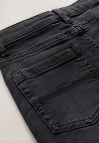 Gros plan d'une poche arrière d'un jean en denim noir avec des coutures visibles et des passants de ceinture sur un fond clair.
