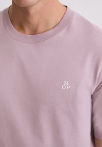 Világoslila, kerek nyakú póló puha anyagból. Kis fehér logó található a mellrészen, sima és egyenletes textúrával.