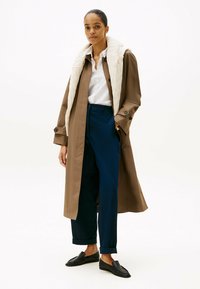 Cappotto trench marrone con colletto trapuntato crema, abbinato a pantaloni blu navy e mocassini neri. L'outfit include una camicia bianca sotto.
