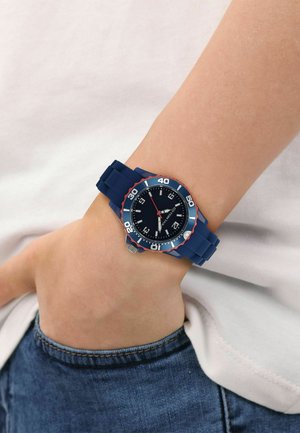 Blue wristwatch with a black dial and red accents worn on a person's left wrist, showing time around 7:40, hand partially in jeans pocket.