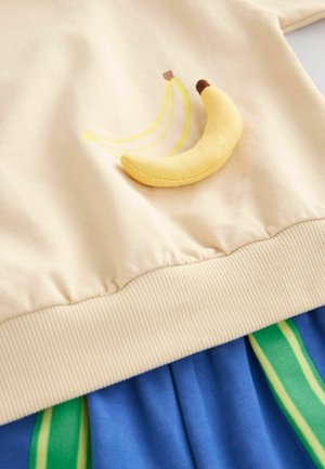 Hellbeige Sweatshirt mit gelbem Bananenmotiv und plüschiger Bananenakzent; kombiniert mit einem blauen Rock mit grünen Streifen. Weicher Baumwollstoff.