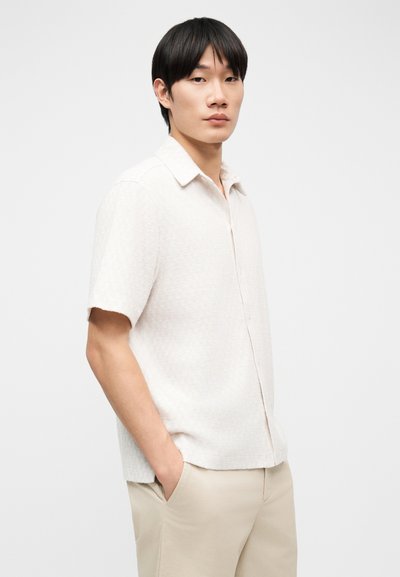 Νέος άνδρας με μαύρα μαλλιά, φορώντας λευκό υφασμάτινο πουκάμισο με κοντά μανίκια και μπεζ παντελόνι, στέκεται με τα χέρια στις τσέπες μπροστά σε λευκό φόντο.