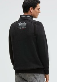 Schwarzer, strukturierter Pullover mit hohem Kragen und gerippten Bündchen. Auf der Rückseite befindet sich ein grafischer Text sowie ein Logo in kontrastierenden Farben. Lockerer Sitz.