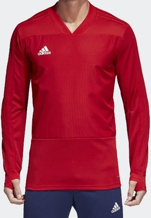 Mężczyzna ubrany w czerwoną sportową bluzę Adidas z długimi rękawami i granatowe spodnie Adidas, stoi z rękami luźno opuszczonymi wzdłuż ciała.