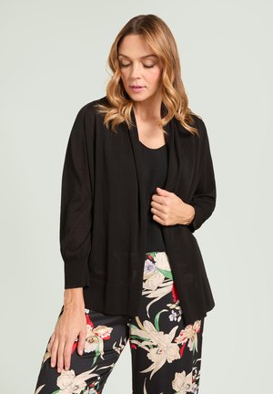 Kvinde med bølget blondt hår iført en sort åben cardigan, sort top og sorte bukser med blomsterprint, der kigger nedad med den ene hånd på cardiganen.