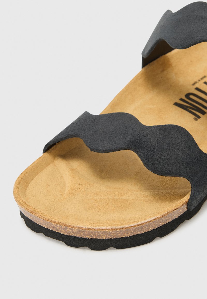 Czarne zamszowe sandały z falistymi paskami, brązowa wyściółka z gumowego podłoża, oraz teksturowana gumowa podeszwa. Posiada zaokrąglone krawędzie i minimalne detale.