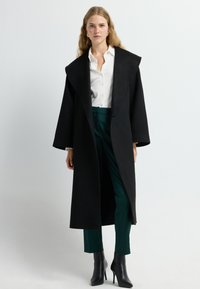 Zwarte wollen jas met een brede kraag en lange mouwen, gecombineerd met een witte button-up shirt en groene broek. Bevat zwarte laarzen met hoge hakken.