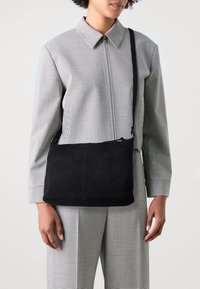 Persona che indossa un giubbotto e pantaloni a scacchi bianco e nero, mentre porta una borsa a tracolla in suede nera senza decorazioni con una cerniera.