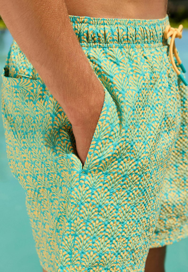 Person trägt türkis-gelbe gemusterte Badeshorts, hat die Hand in der Seitentasche und steht in der Nähe eines Pools.