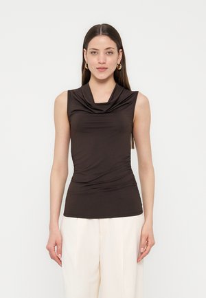 Kobieta z długimi brązowymi włosami, nosząca złote kolczyki kółka, czarny, bezrękawnikowy top z marszczonym dekoltem oraz białe, szerokie spodnie z wysokim stanem, zwrócona twarzą do przodu.