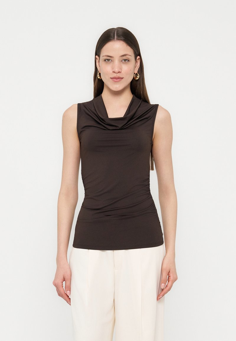 Γυναίκα με μακριά καστανά μαλλιά, που φοράει χρυσούς κρίκους στα αυτιά, μαύρο αμάνικο τοπ με ντεκολτέ και λευκό φαρδύ παντελόνι με ψηλή μέση, κοιτάζοντας μπροστά.