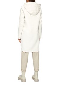 Cappotto bianco lungo con cappuccio imbottito, texture liscia, taglio dritto, abbinato a pantaloni beigi roll-up e stivali chiari.