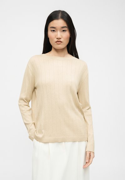 Femme aux cheveux bruns raides portant un pull côtelé beige et une jupe blanche, debout avec une main dans la poche devant un fond blanc.