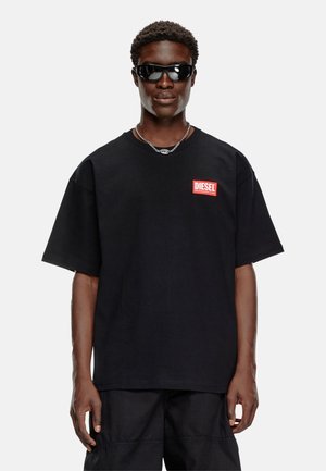 Schwarzes Baumwoll-T-Shirt mit kurzen Ärmeln, versehen mit einem roten "DIESEL"-Logo auf der linken Brust. Getragen mit dunklen Sonnenbrillen und einer Kettenkette.