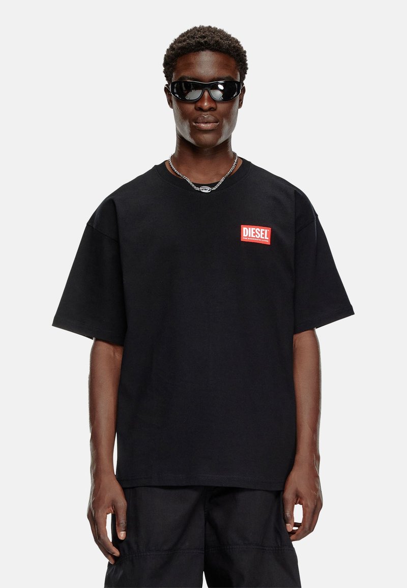 Sort bomuldst-shirt med korte ærmer, der har et rødt "DIESEL" logo på venstre bryst. Iført mørke solbriller og en kæde-halskæde.