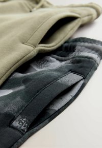 Hanorac verde cu o textură moale; prezintă căptușeală de camuflaj în nuanțe de gri închis și negru, evidențiind modele și materiale contrastante.