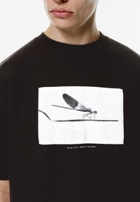 Sort bomulds T-shirt med et hvidt rektangulært grafisk motiv af en guldsmed, der sidder på en streg. Teksten nedenunder siger "RADIKALE NYE VISIONS."