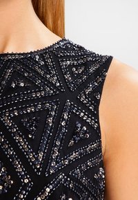 Närbild av en ärmlös svart klänning med axel som har geometriska mönster av glänsande svarta och silvriga paljetter och pärlor.