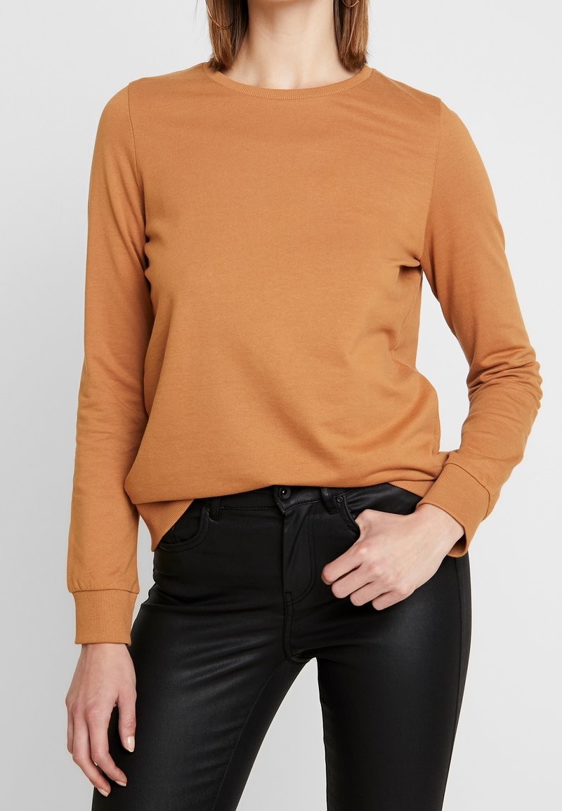 Femme portant un pull à manches longues couleur camel et un pantalon en cuir noir ajusté, une main dans la poche, sur un fond uni.