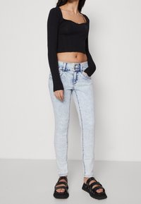 Žena v černém dlouhém topu s krátkým střihem, světlých modrých skinny džínách s vysokým pasem a dvojitým knoflíkem, a černých platformových sandálech.