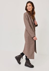 Barna gyapjú keverék hosszú kabát, hegyes hajtókákkal, oldalsó zsebekkel és laza szabással, fekete fűzős csizmával és nadrággal párosítva.