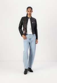 Giubbotto in pelle nera con un design strutturato, t-shirt bianca, jeans azzurri chiari e stivaletti neri alla caviglia. Una borsa a tracolla marrone aggiunge contrasto.