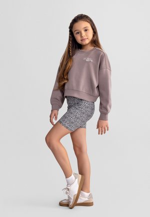 Mladé dievča s dlhými vlasmi, oblečené v fialovom mikine, kraťasoch so vzorom leopardieho kožušiny, bielych ponožkách a bielych teniskách, stojace pred jednoliatym pozadím.