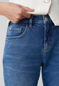 Jeans en denim bleu avec un design ajusté, présentant une fermeture à bouton à l'avant et des coutures contrastées. Comprend une poche et une texture lisse.