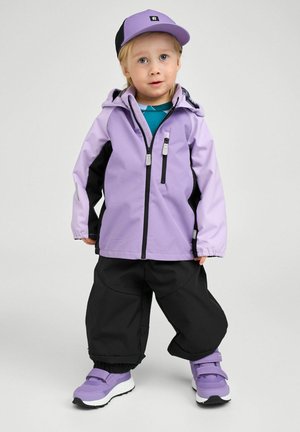 Małe dziecko w fioletowej kurtce, dopasowanej czapce i butach, czarnych spodniach, stojące na tle jednolitego jasnego tła.