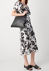 Sac fourre-tout en cuir noir au toucher lisse, avec un design simple. Le sac est porté avec une robe florale noir et blanc.